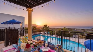 A poolside setting with a sunset view, a table with food and drinks, and a pergola with string lights. - Pool Villa Overlooking the Sea (Rhodes)