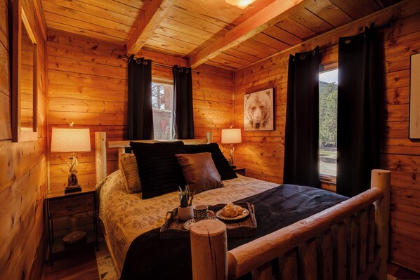 Un dormitorio de cabaña de madera con una cama, dos lámparas de noche, un cuadro enmarco y una ventana con cortinas.