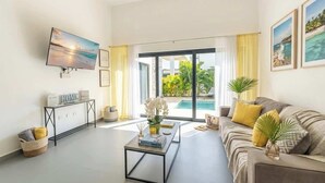 A modern living room with a flat-screen TV, a sofa, a coffee table, and a view of a pool and greenery. - Private Pool Villa in Sosua's Ocean Village (Sosúa)