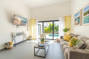 A modern living room with a flat-screen TV, a sofa, a coffee table, and a view of a pool and greenery. - Modern Luxury Villa with Private Pool Ocean Village (Sosúa)