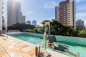 Ein Pool auf dem Dach mit einer Holzterrasse, einer Liege und Blick auf umliegende Hochhäuser.