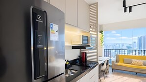 A modern kitchen with a stainless steel refrigerator, a microwave, and a coffee maker. There is a dining area with a yellow sofa and a view of a cityscape through large windows. - Smart Bourbon 2009 (São Paulo)