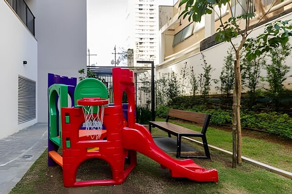 Ein Spielplatz mit Rutsche und Klettergerüsten, einer Bank und einem kleinen Baum in einer Stadtumgebung.