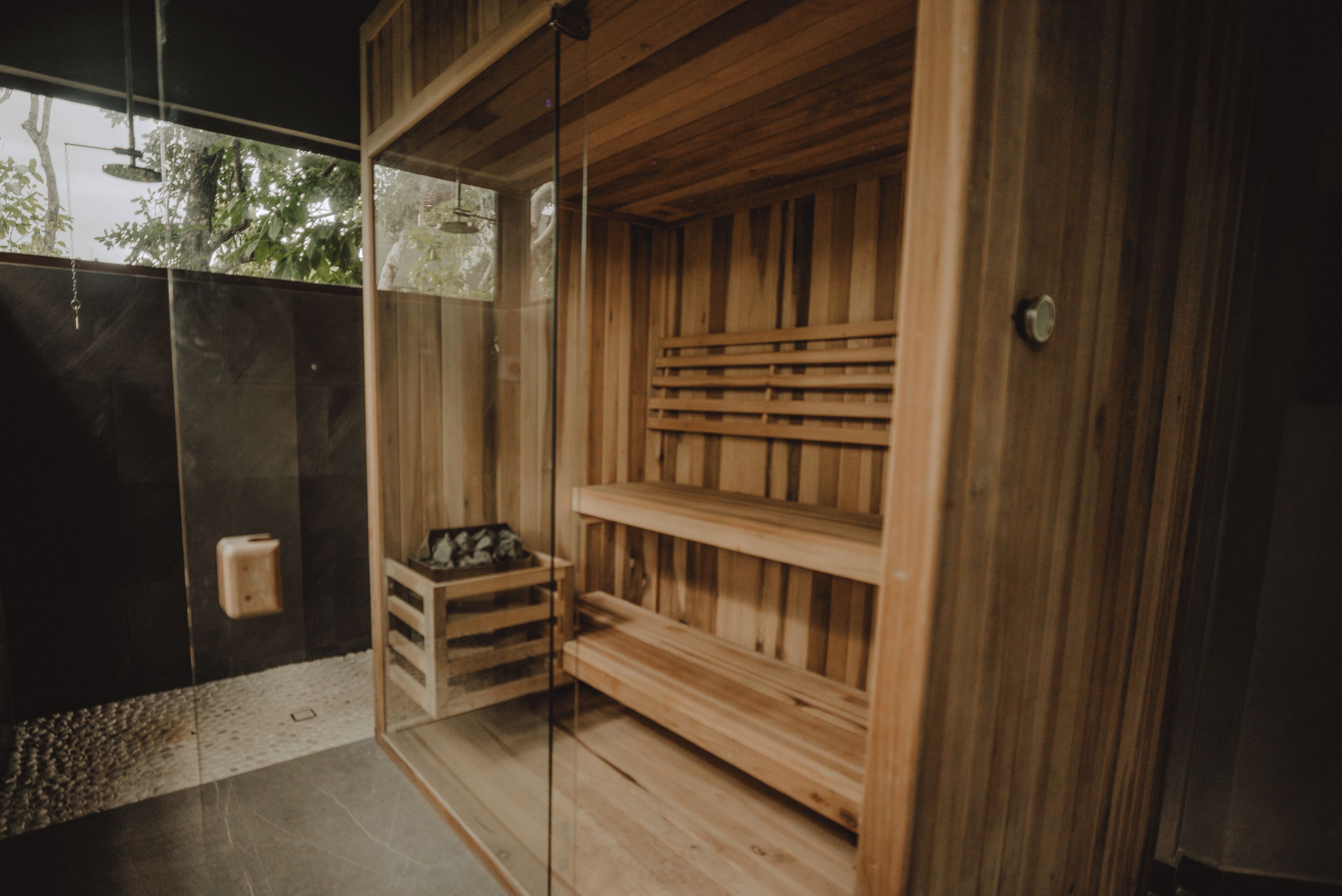 A wooden sauna with benches and a glass door, a towel dispenser, and a small wooden stool.