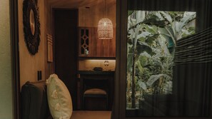 A room with a wooden cabinet, a hanging light fixture, a small table with a glass, and a view of a garden with banana plants. - Private Jungle Retreat + Fast Wi-Fi & Private Pool (Tulum)