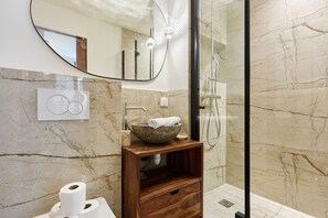 Un bagno con parete in marmo, mobile lavabo in legno, lavabo tondo, specchio e cabina doccia in vetro.