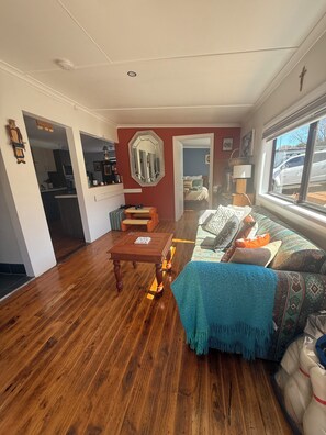 A living room with a wooden floor, a sofa with patterned cushions, a coffee table, and a mirror on the wall.