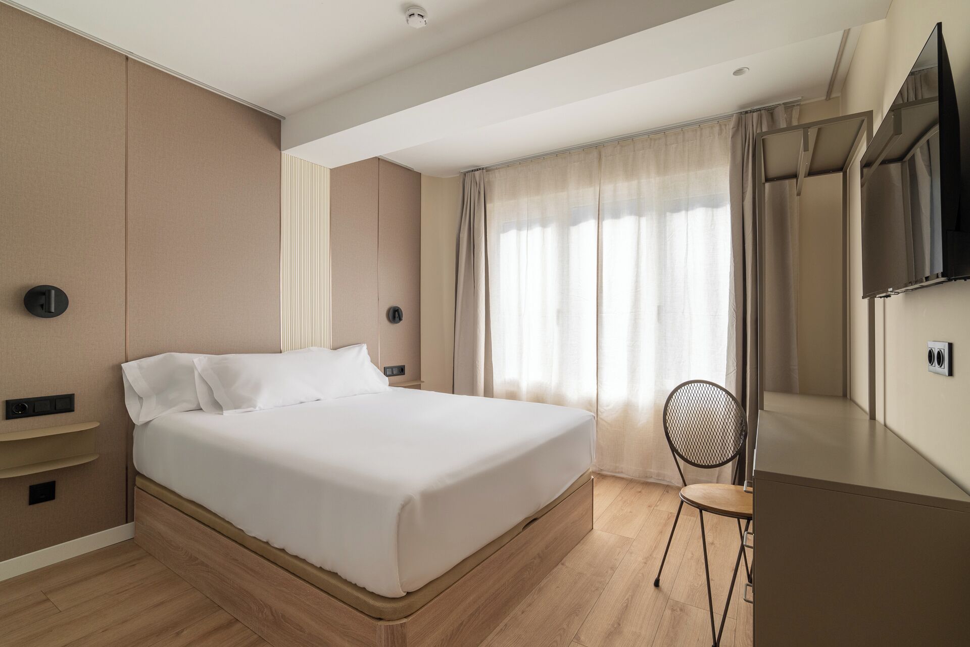 Una habitación de hotel con una cama, una silla, un televisor y un ventanal con cortinas.