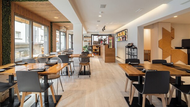 Un restaurante moderno con mesas y sillas de madera, una recepción y vistas a la calle a través de amplios ventanales.