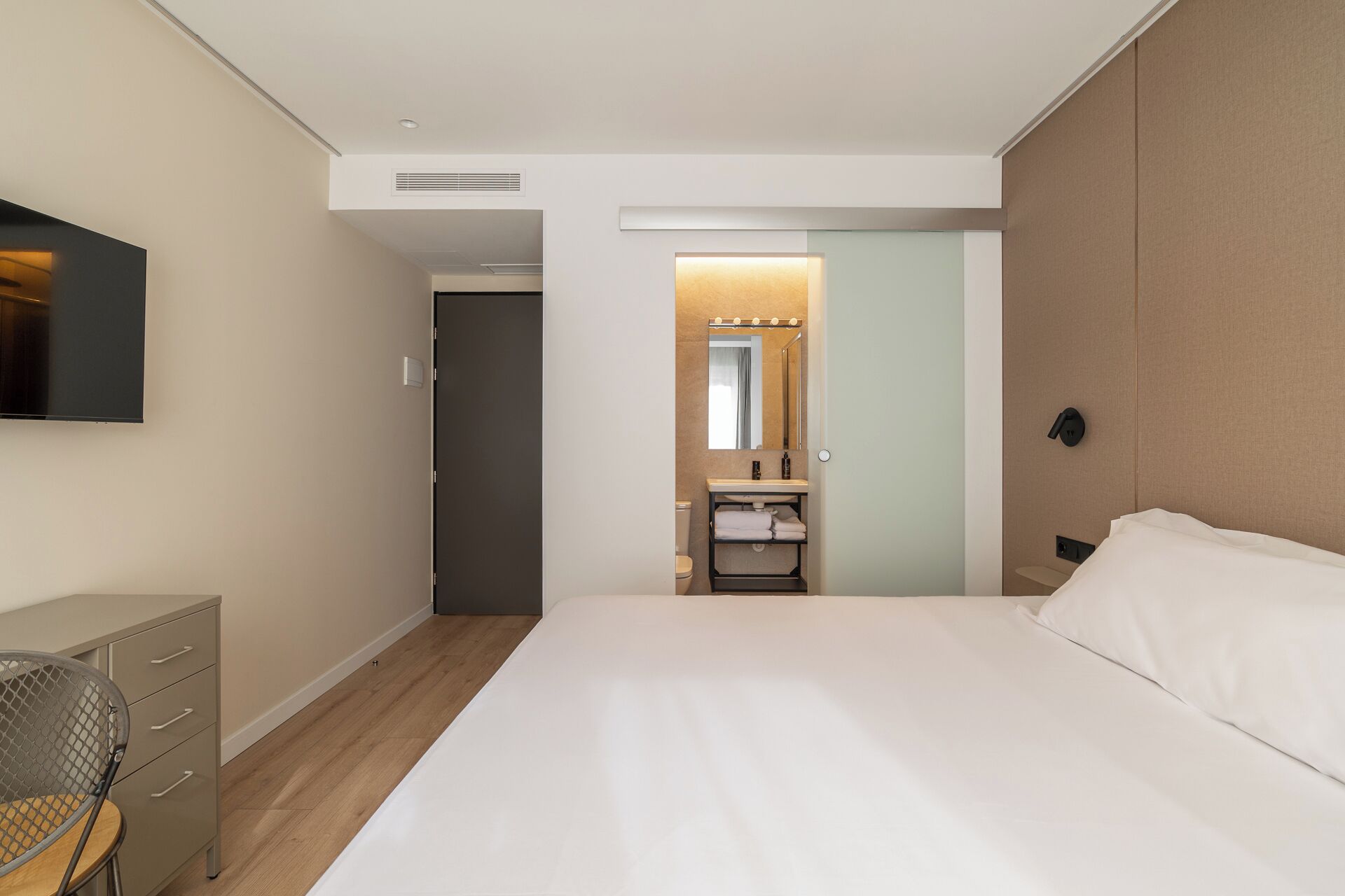 A hotel room with a bed, a desk, a chair, a bathroom with a sink and mirror, and a television on the wall.