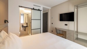 Habitación de hotel con cama, escritorio con silla, televisor, baño con espejo y lavamanos, y puerta corredera.