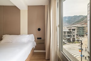 Habitación de hotel con cama, panel de control en la pared y ventana con vista a un paisaje urbano.