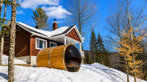 Una cabaña con un barril de madera, tejado cubierto de nieve y un cielo azul despejado.