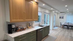 A modern kitchen with wooden cabinets, a coffee station, and a dining area with a table and chairs. - Otter Cove Cottage, completely renovated house on Campobello (Wilsons Beach)