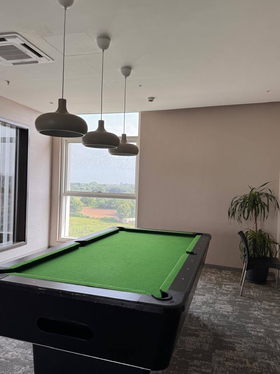 Una mesa de billar en una habitación con un ventanal grande, lámparas colgantes y una planta en maceta.