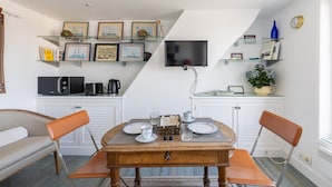 Een eetgedeelte met een houten tafel, twee stoelen, een aan de muur bevestigde televisie en planken met ingelijste foto's en decoratieve objecten.