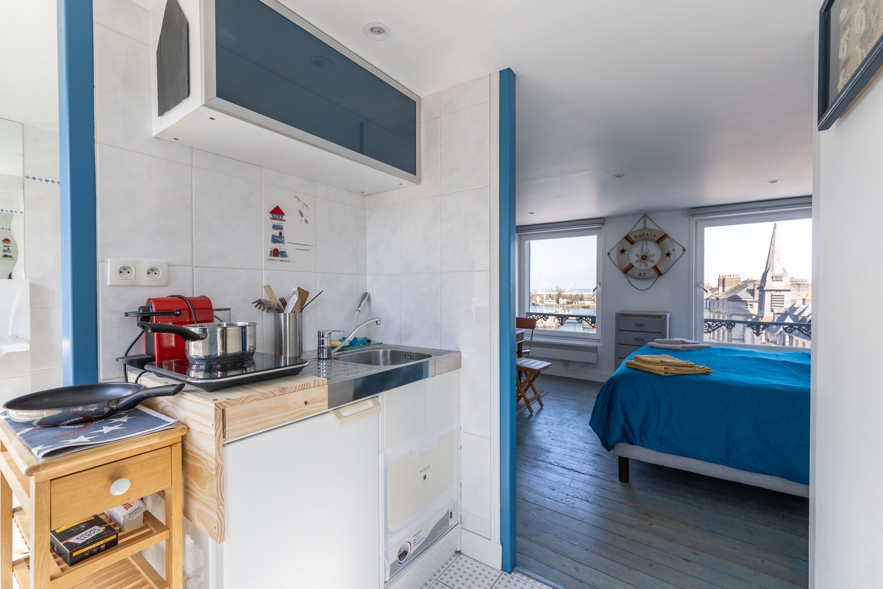 Une pièce compacte dotée d’une kitchenette, d’un lit recouvert d’une courtepointe bleue et offrant une vue sur la ville grâce à de grandes fenêtres.