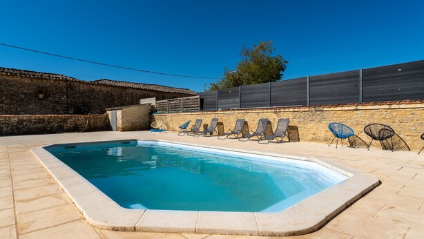 Et rektangulært swimmingpool med flisebelagt solterrasse, omgivet af liggestole og en stenvæg.