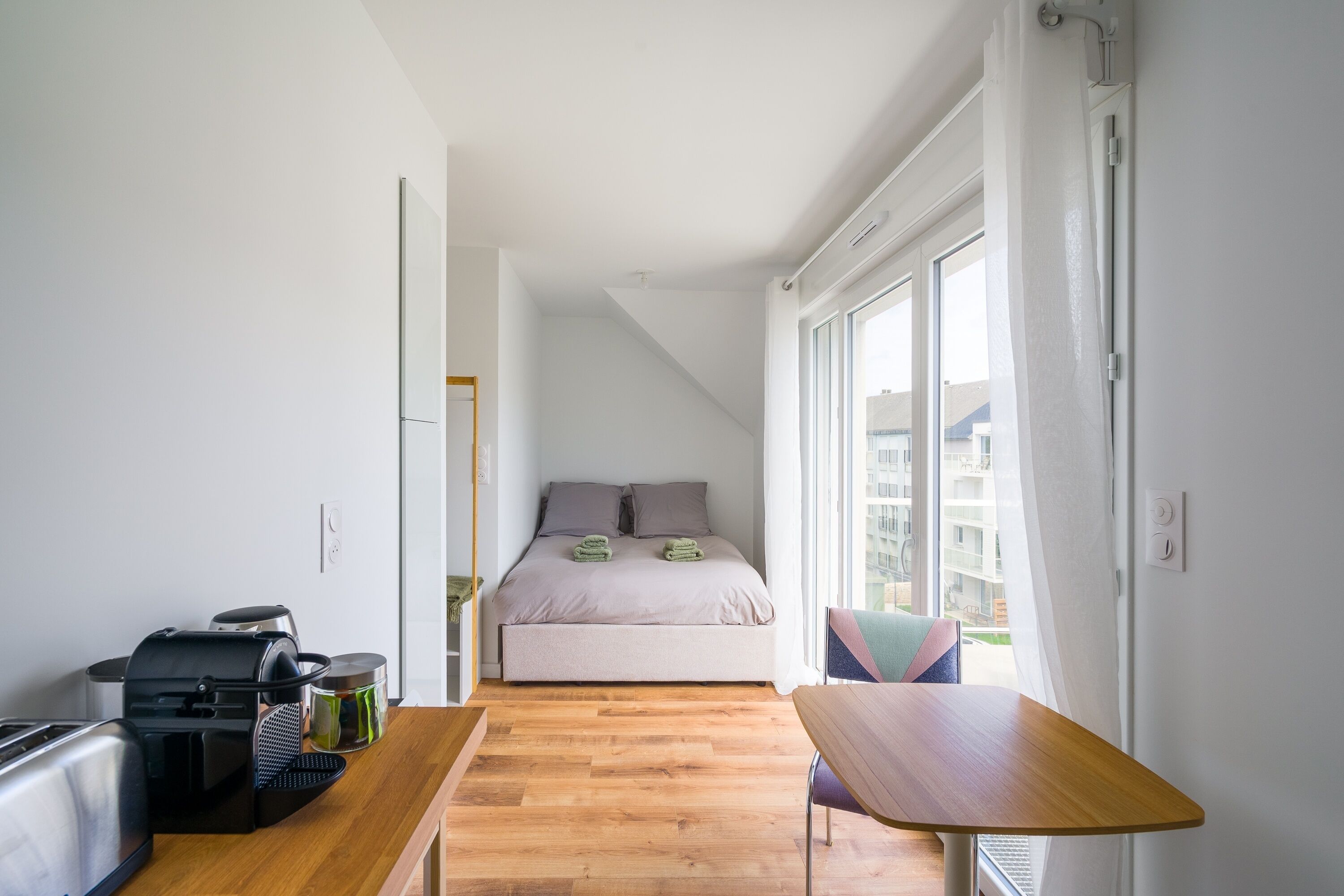A bedroom with a bed, a window, a table, and a kitchenette.