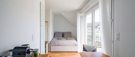 A bedroom with a bed, a window, a table, and a kitchenette.