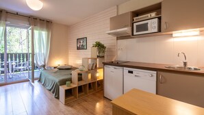 A compact kitchen with a microwave, sink, and dishwasher. A bed with a green bedspread is adjacent to the kitchen, and a balcony with a view of trees is visible. - La Bulle d’Ax - Domaine de la Vallée d'Ax (Ax-les-Thermes)