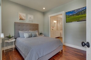 Ein Schlafzimmer mit einem Bett, einem Nachttisch, einer Topfpflanze, einem an der Wand montierten Fernseher und einem durchgehenden Türspalt sichtbaren Badezimmer.