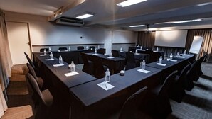 Un salón de conferencias con largas mesas, sillas, botellas de agua y una pantalla para proyector.