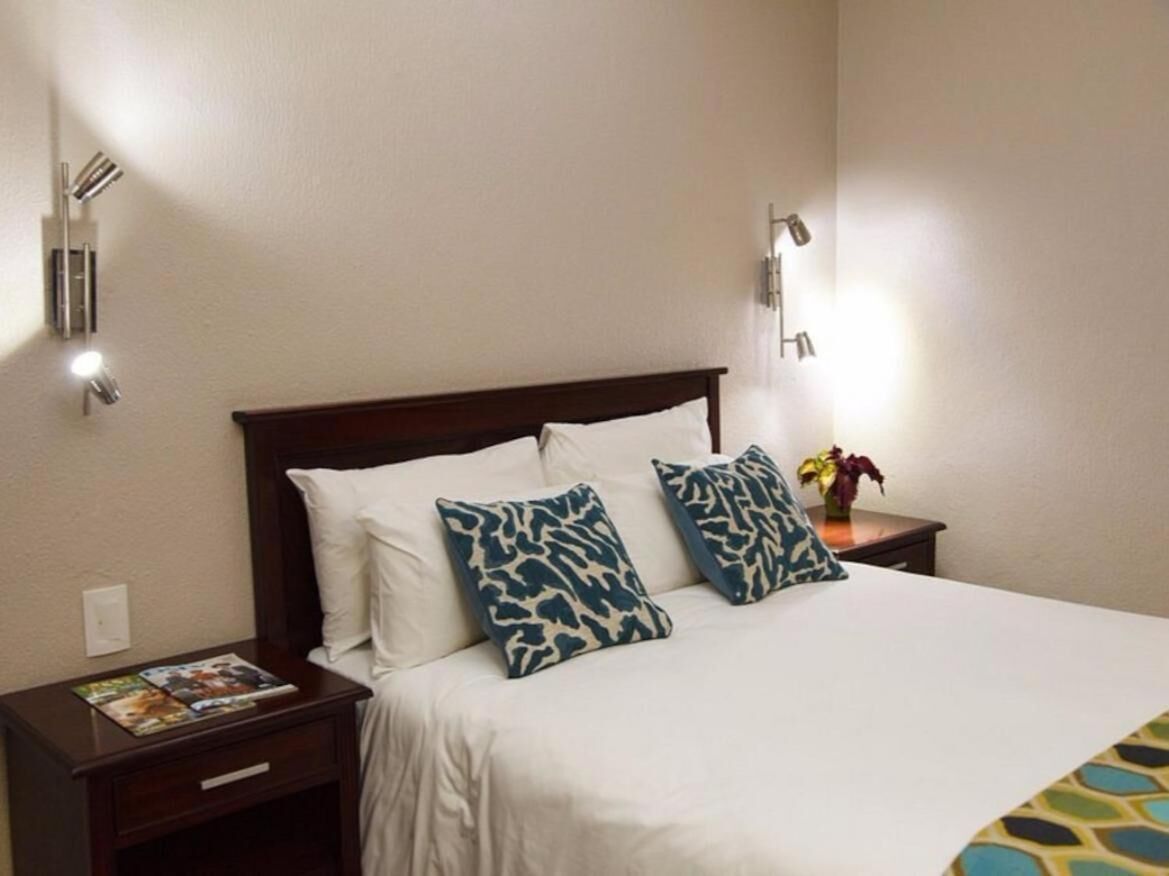 A hotel room with a bed, two pillows, a nightstand, a lamp, and a small table with a vase.
