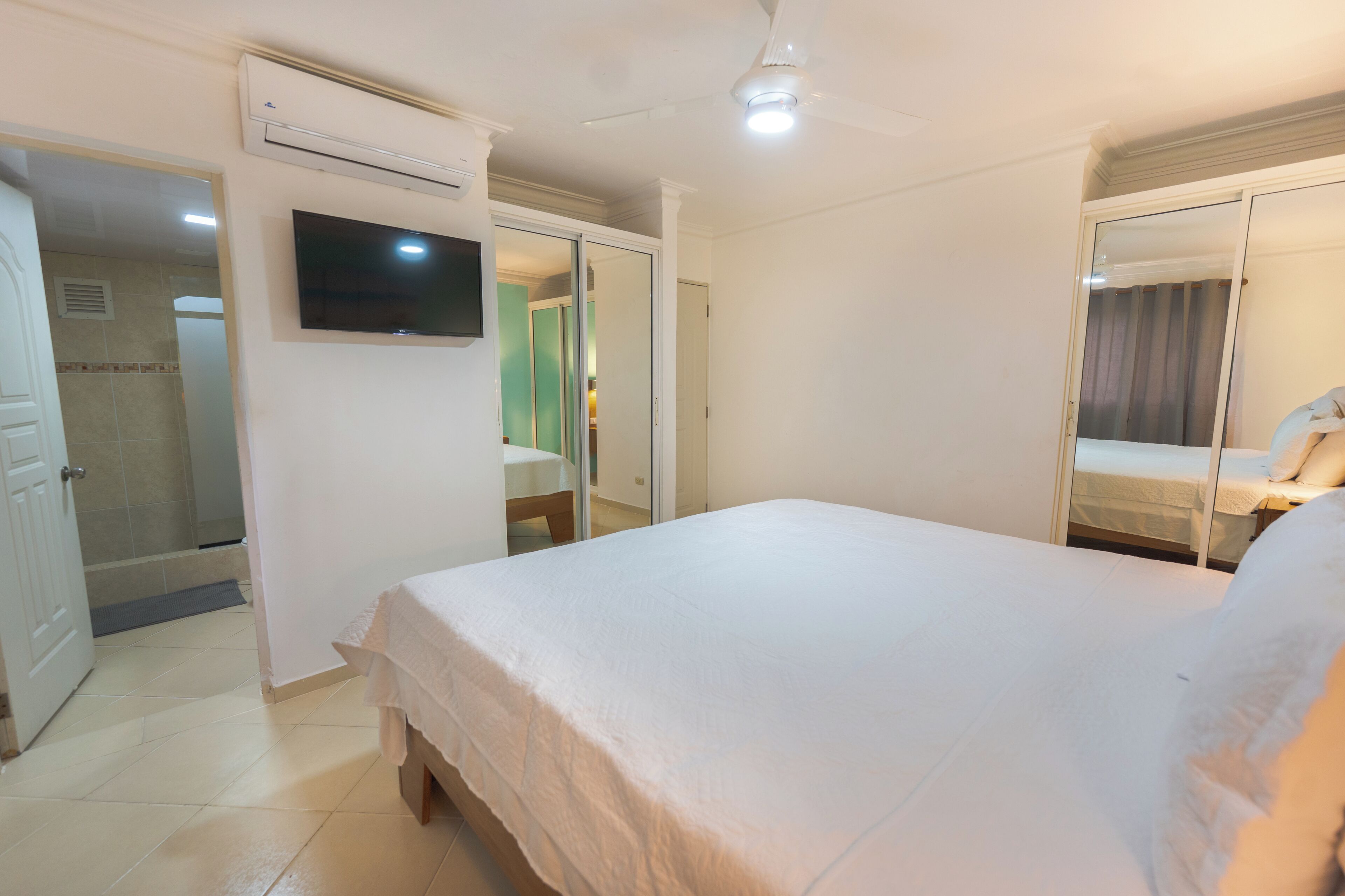 A bedroom with a bed, a TV mounted on the wall, an air conditioning unit, and a ceiling fan.