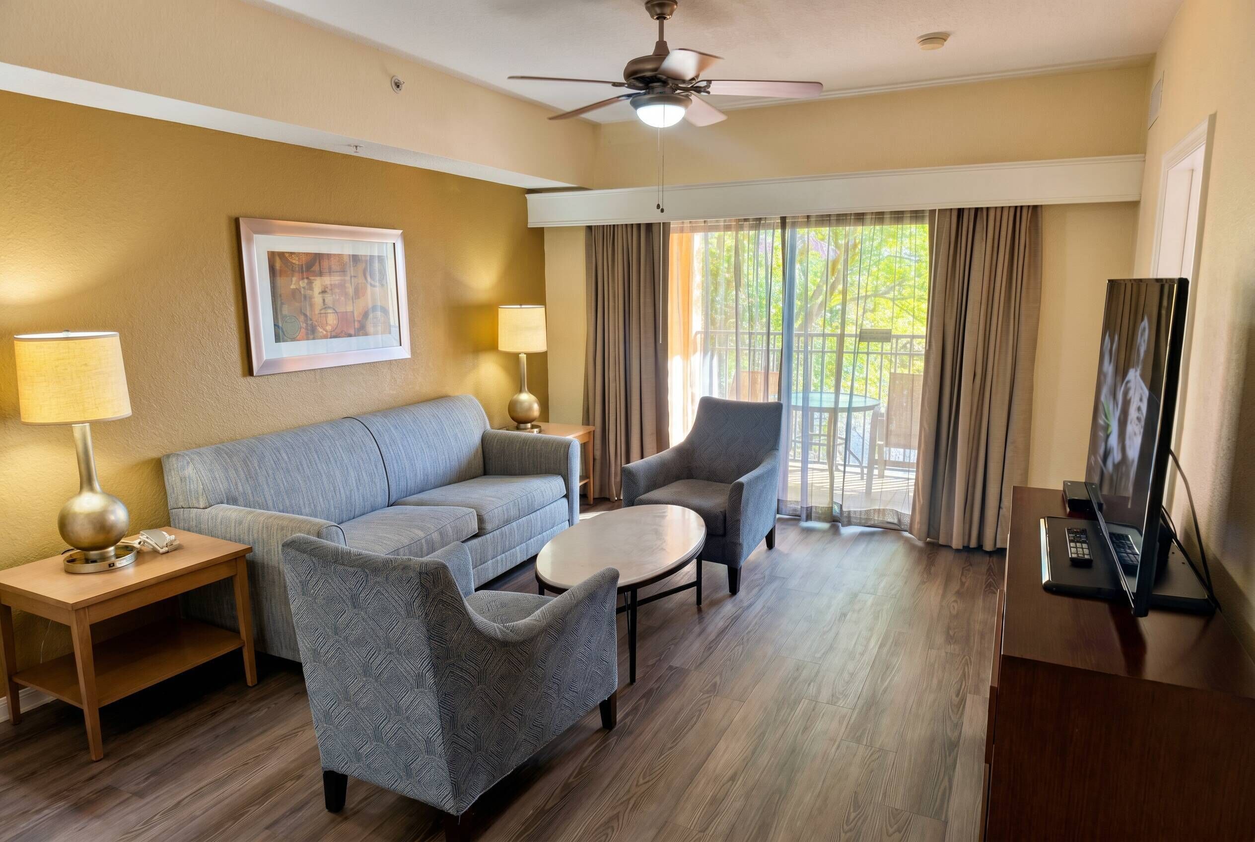 Habitación de hotel con sofá, sillón, mesa redonda, escritorio con computadora y ventilador de techo.