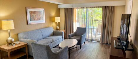 Habitación de hotel con sofá, sillón, mesa redonda, escritorio con computadora y ventilador de techo.