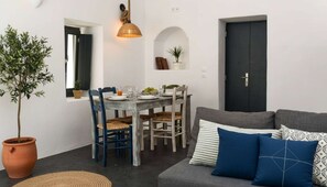 A dining area with a table set for two, a sofa with patterned cushions, and a potted plant. - Grand Village Stay for Seven (Santorini)