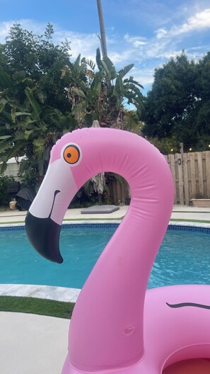 Ein rosa Flamingo-Schwimmpfad in einem Garten mit Pool, umgeben von Grünpflanzen und einem Holzzaun.