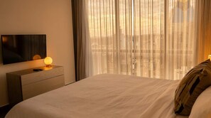 Hotellihuone, jossa on sänky, yöpöytä lampun kanssa, seinälle asennettu taulutelevisio ja näkymä kaupunkiin ohuiden verhojen läpi.