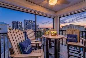 A balcony with wooden chairs, a table, and a view of buildings at dusk. - Beachfront Escape with Pool, Tiki Huts & Stunning Gulf Views - Casa Bleu - Roelens (Fort Myers Beach)