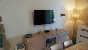 A modern room with a flat-screen TV mounted on the wall, a wooden cabinet with drawers, a dining table with chairs, and a small trash bin.