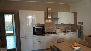 A modern kitchen with white cabinets, a gas stove, an oven, and a sink. There is a dining table with chairs and a bowl of fruit on it.