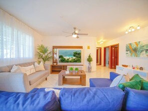 A modern living room with a flat-screen TV, a ceiling fan, and a large window with sheer curtains. - Cozy 7BR Villa BIG Pool Close to Beach! (Sosúa)