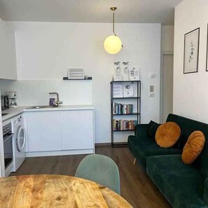 Ein kompakter Wohnbereich mit Küchenzeile, Essplatz, einem grünen Sofa mit orangefarbenen Kissen und einem Bücherregal.