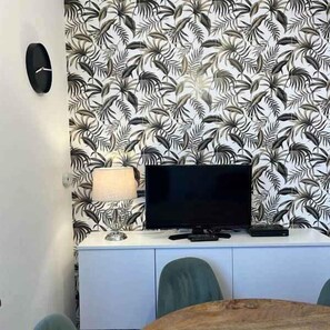 Ein Zimmer mit Fernseher auf einer weißen Kommode, einer Lampe und einem Tisch mit Stühlen.