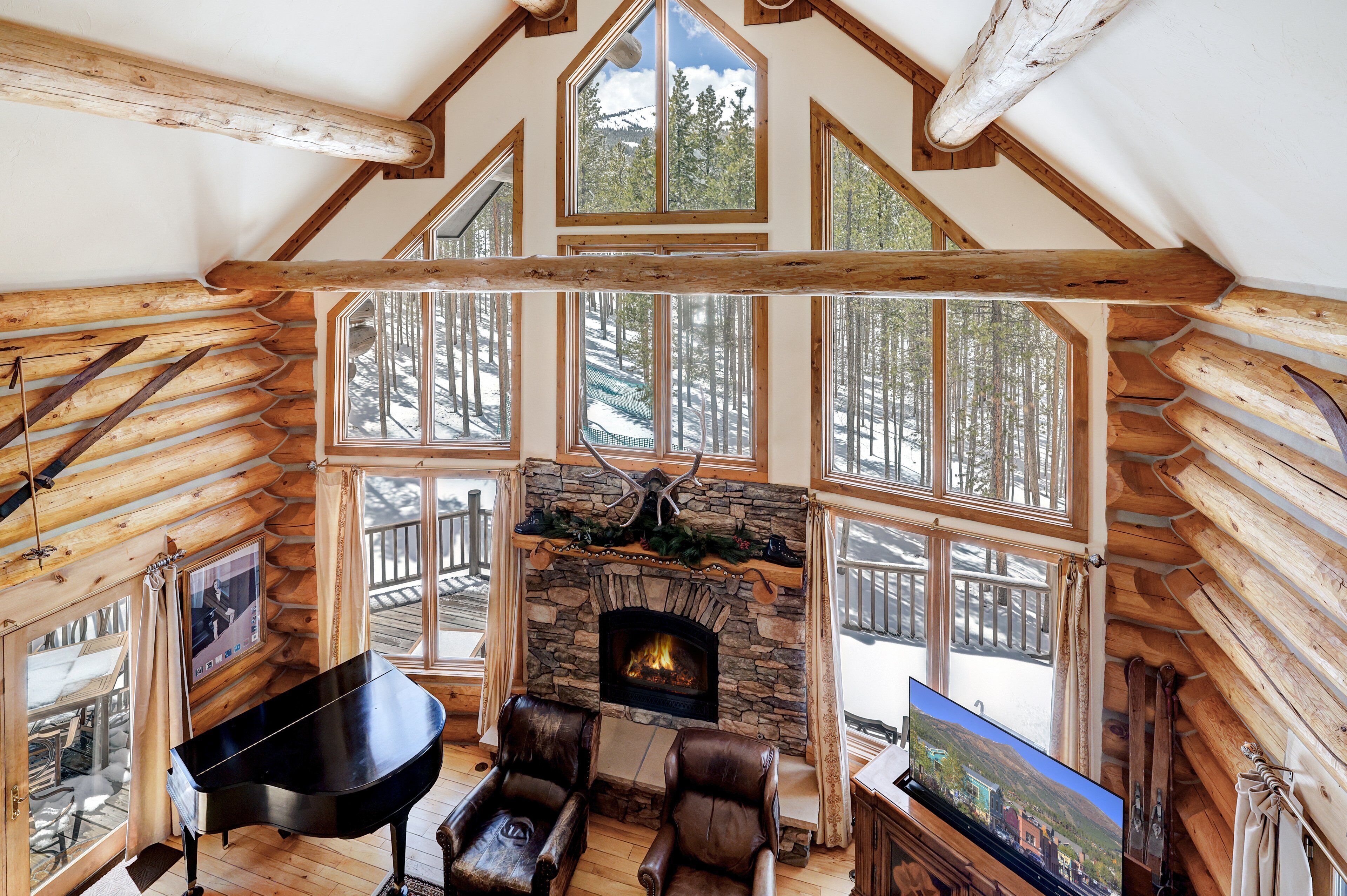 A log cabin interior with a stone fireplace, wooden beams, large windows, a piano, and leather chairs.