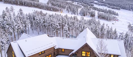 A log cabin with multiple windows and a garage, surrounded by snow-covered trees and a mountainous backdrop.