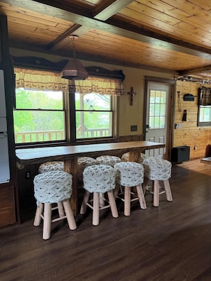 A wooden interior with a bar area, cushioned stools, and a view of greenery outside. - Spaulding Lake Cottage: 4-acre lakefront retreat in Northern Maine (Oakfield)