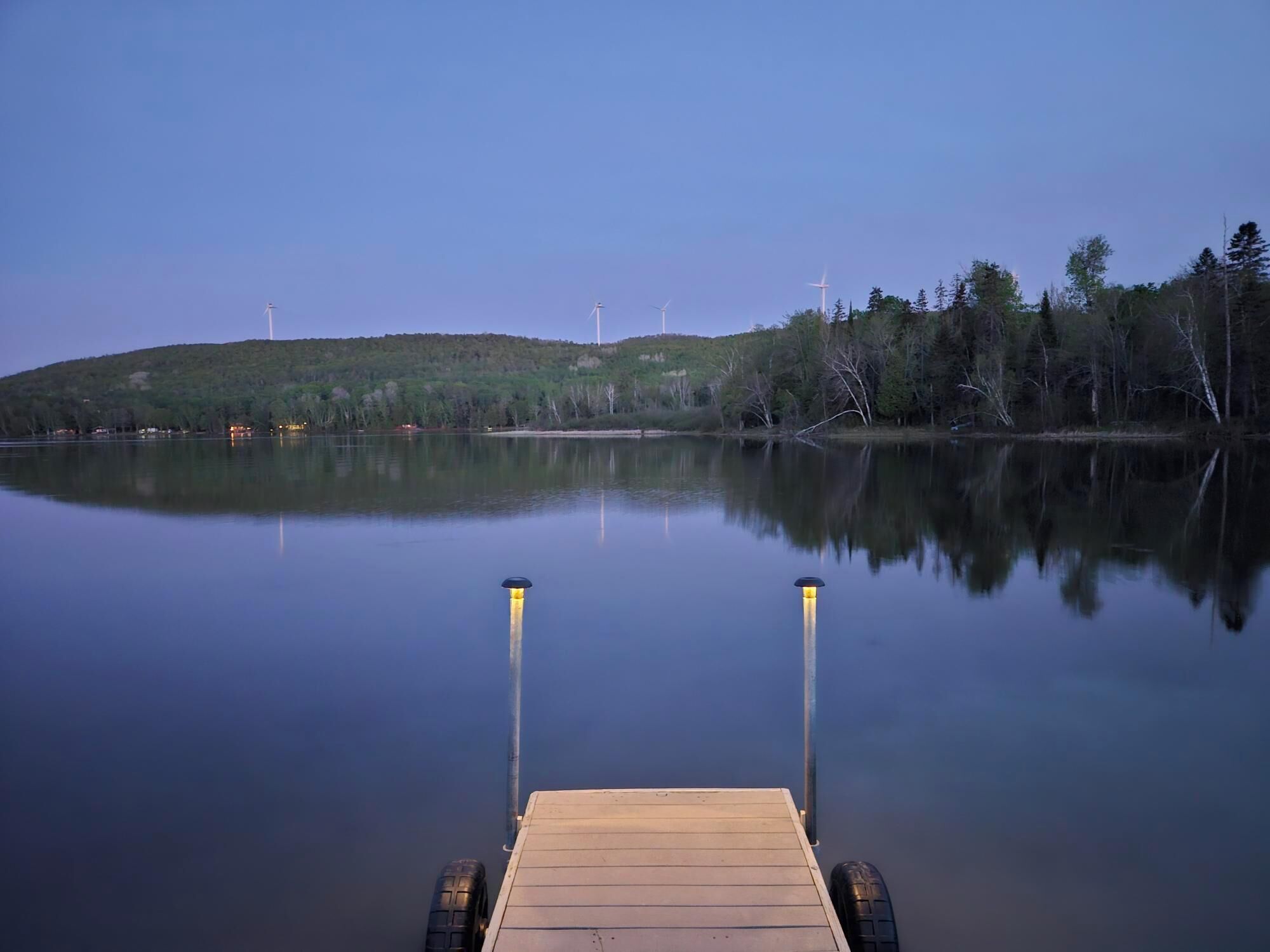 A calm lake with a wooden dock extending into the water, surrounded by trees and hills.