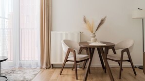 Uma sala de jantar com uma mesa redonda, duas cadeiras, um vaso com plantas secas, um radiador e uma janela com cortinas leves.