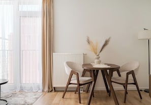 A dining area with a round table, two chairs, a vase with dried plants, a radiator, and a window with sheer curtains. - Browary Warszawskie Harmony Studio (Warszawa)