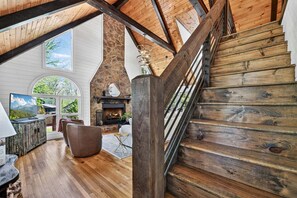 A wooden staircase leading to a loft area with a fireplace, a television, and a stone wall. - Laurel Ridge Retreat - Hot Tub - Wood-burning Fireplace - Fire Pit (Morganton)