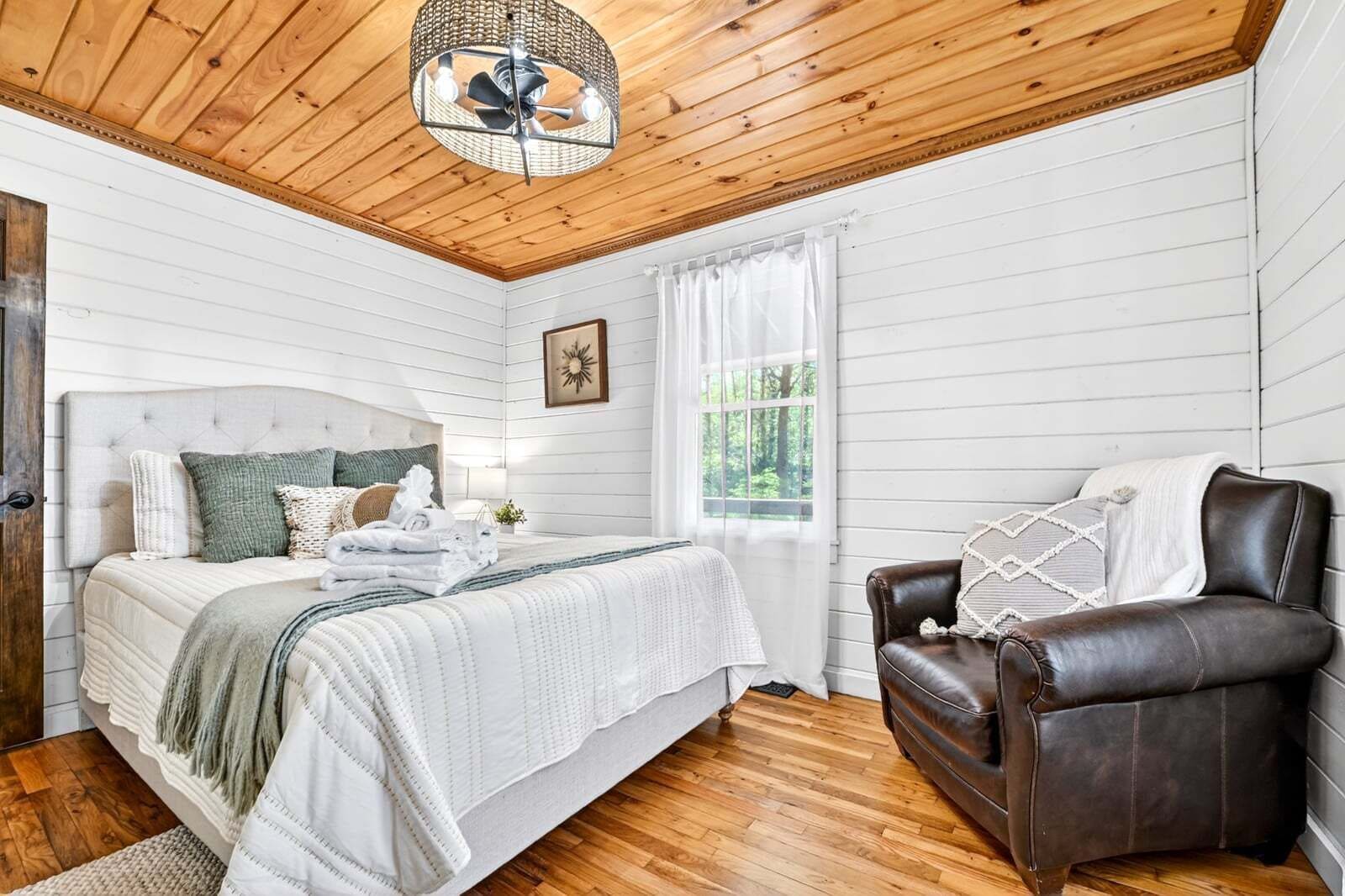 A bedroom with a bed, a leather chair, a wooden ceiling, and a window.
