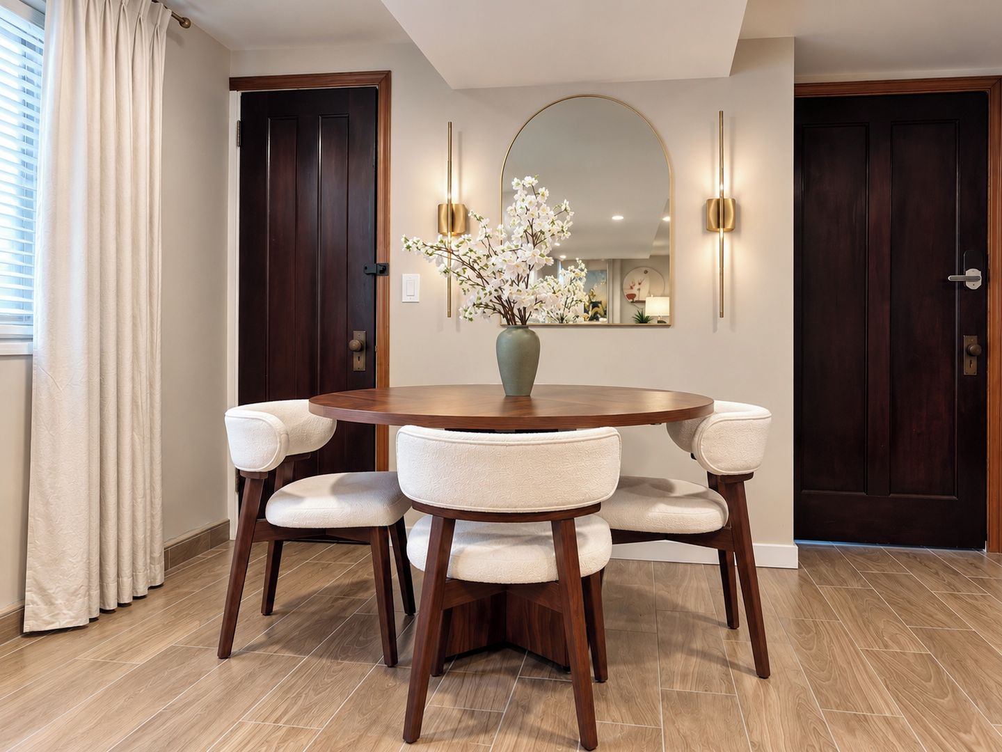 Un coin repas comprenant une table ronde en bois, quatre chaises rembourrées, un vase avec des fleurs blanches et deux appliques murales.