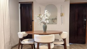 Un comedor con una mesa redonda de madera, cuatro sillas blancas tapizadas, un jarrón con flores blancas, un espejo y dos puertas.